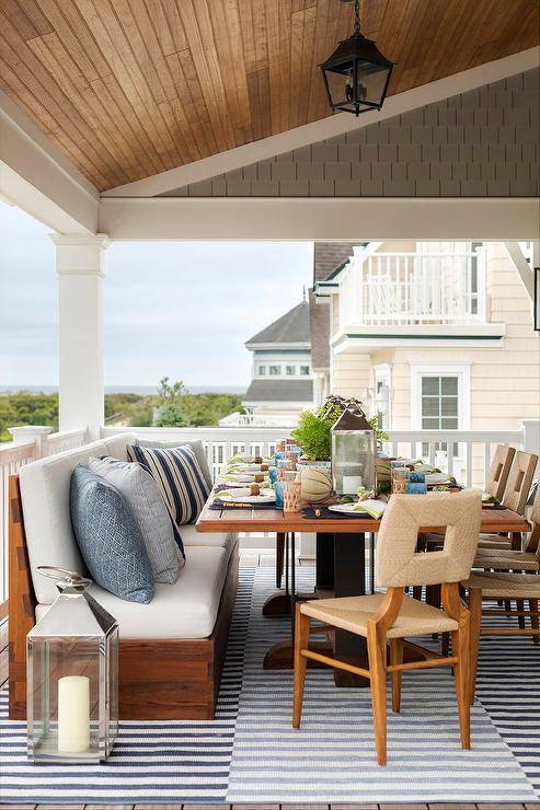 Abaca dining chairs sit on a covered deck on a black and white striped rug placed beneath a teak slatted dining table paired with a teak dining bench complemented with white and blue pillows. A black lantern hangs over the table from a brown plank ceiling.