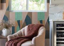 Earth toned geometric wallpaper frames a window located over a mauve tufted accent chair finished with wooden legs and draped with a brown throw blanket.