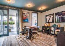 Well appointed game room features a round game table surrounded by wood and brass industrial chairs placed on a beige striped rug under a blue painted ceiling lined with blue crown molding and fitted with star flush mounts. Framed sports jerseys hang from a light gray wall above an industrial cabinet as a framed movie print is fixed beside a blue glass front French patio door covered in sheer beige pleated curtains.