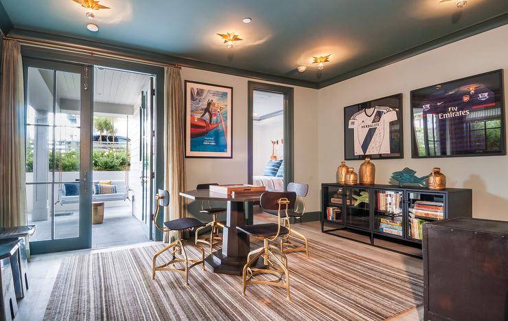 Well appointed game room features a round game table surrounded by wood and brass industrial chairs placed on a beige striped rug under a blue painted ceiling lined with blue crown molding and fitted with star flush mounts. Framed sports jerseys hang from a light gray wall above an industrial cabinet as a framed movie print is fixed beside a blue glass front French patio door covered in sheer beige pleated curtains.