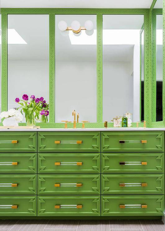 A bright green ornate washstand adorned with brass and glass pulls is finished with a brass faucet fixed beneath a green Greek key mirror.