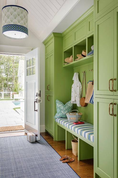 A blue and green striped cushion accents a built-in bright green mudroom bench fixed against green shiplap trim fitted with brass coat hooks mounted under green cubbies. The bench is flanked by stacked green cabinetry, while a blue bound sisal rug leads under a blue and green drum pendant to a white front door with six glass panels.