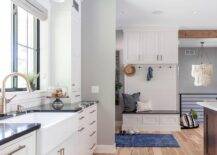 A built-in small mudroom bench with dark blue and white pillows under built-in cabinets on white shiplap trim and a blue over-dyed rug are next to a black and white kitchen.