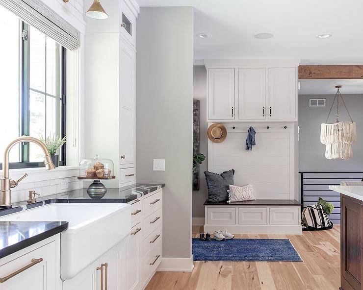 A built-in small mudroom bench with dark blue and white pillows under built-in cabinets on white shiplap trim and a blue over-dyed rug are next to a black and white kitchen.