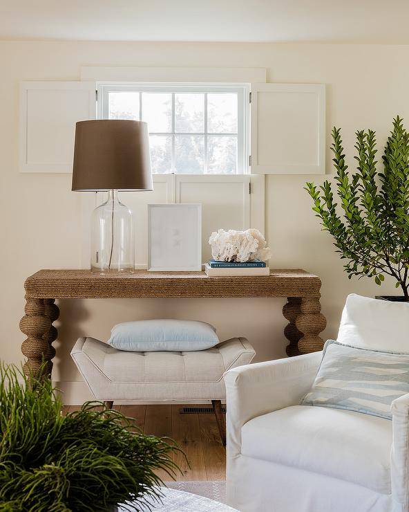 Anthropologie Jute Wrapped Console Table surely makes a statement in a cottage living room showcasing a glass lamp with a brown shade and a cream tufted bench underneath. Indoor plants white window trim and white surrounding furnishings create a dwelling sure to appeal the decorative eye and offer a place to rest and unwind.