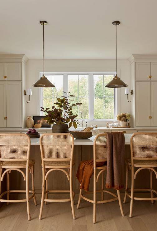 Vintage cane stools are placed at a brown oak island topped with a marble countertop lit by vintage brass lanterns.