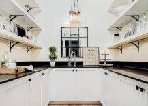 Black and white U-shaped cottage style pantry with white vertical shiplap walls boasts white shelves with brass brackets and custom shelf lighting, white cupboards with oil-rubbed bronze square knobs, lit by a vintage glass bottle chandelier.