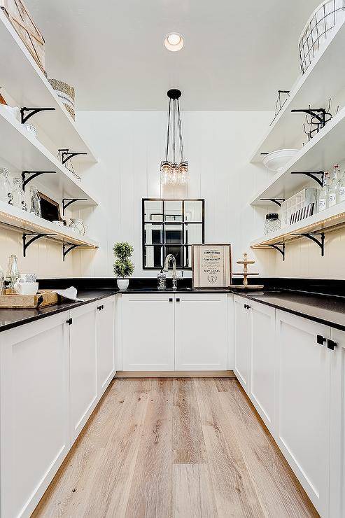 Black and white U-shaped cottage style pantry with white vertical shiplap walls boasts white shelves with brass brackets and custom shelf lighting, white cupboards with oil-rubbed bronze square knobs, lit by a vintage glass bottle chandelier.