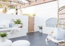 Backyard oasis features a custom water fountain and string lights lighting a pergola fitted with an extra large rattan hanging chair. The chair hangs facing a white built-in sofa topped with a light gray cushion white and gray pillows.