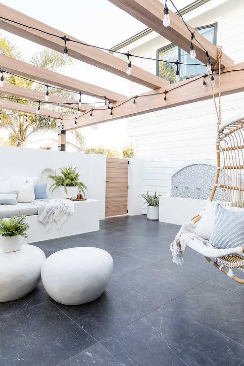 Backyard oasis features a custom water fountain and string lights lighting a pergola fitted with an extra large rattan hanging chair. The chair hangs facing a white built-in sofa topped with a light gray cushion white and gray pillows.