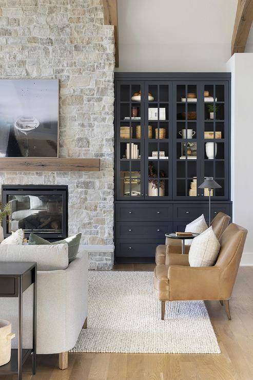 Black built-in display cabinets are fixed in a country style living room beside a gray stone fireplace finished witha hearth and a chunky brown wooden mantel mounted beneath an art piece.