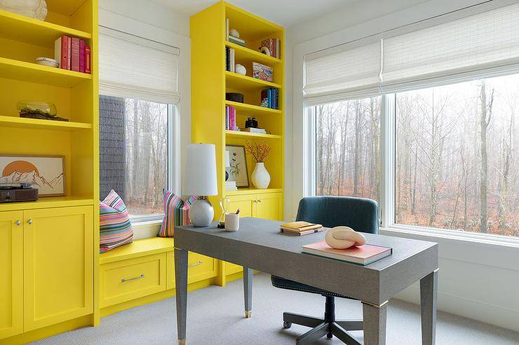 Charming yellow and gray office boasts a gray shagreen desk placed in front of windows covered in white roman shades. Tall yellow bookcases flank a yellow window seat finished wit drawers and topped with a striped pillows.