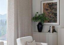 Beige pleated curtains hang behind white boucle chairs complemented with black and white pillows and placed on a white rug flanking a round black accent table.