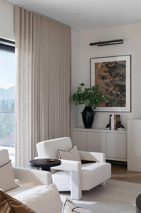 Beige pleated curtains hang behind white boucle chairs complemented with black and white pillows and placed on a white rug flanking a round black accent table.