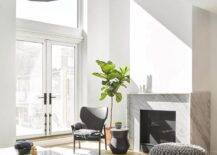 Chic modern living room boasts gray knitted stools placed on a gray rug in front of a modern gray marble fireplace, as a black accent table is paired iwth a black leather chair located in front of a fiddle leaf fig.