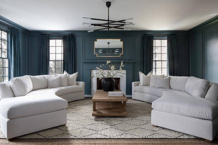 A raffia coffee table sits on a lattice rug between gorgeous curved white sofas lit by a black and silver chandelier. A gold framed mirror hangs from a blue wall with blue trim over a blue fireplace mantel framing a marble surround. Windows are dressed in blue curtains.