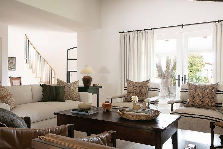 French patio doors covered in white curtains are located behind black and white burlap accent chairs placed facing a brown farmhouse coffee table.