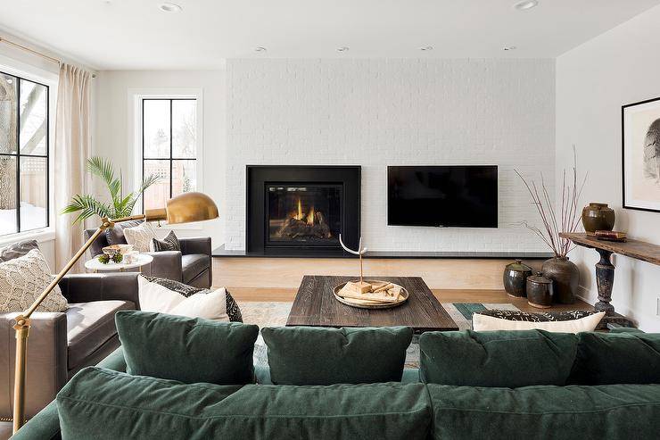 TV mounted next to a fireplace on a white brick wall in a country living room furnished with a hunter green sectional and a brown stained oak coffee table. This cabin home invites a luxurious appeal mixed with rustic wood finishings and brown leather materials.