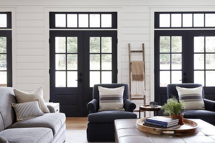 Charming modern farmhouse living room boasts dark blue roll armchairs accented with vintage burlap French pillows facing a large brown leather ottoman. Shiplap walls and black French doors bring a modern farm style finish decorated with a wood throw wall leaning blanket ladder.