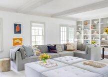 Gorgeous gray living room features a low gray tufted modular sectional placed beside a tree stump end table facing four gray tufted modular ottomans. Abstract blue and orange art hangs beside the sofa and between windows, while styled white built-in cabinets are fixed into white walls lined with light gray trim.