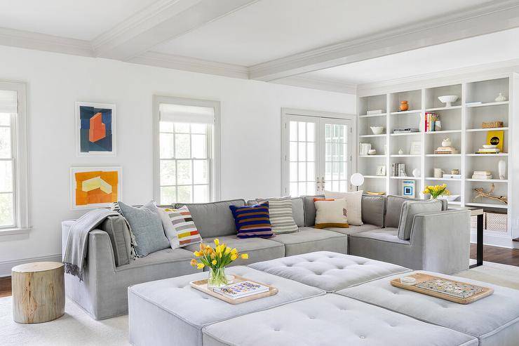 Gorgeous gray living room features a low gray tufted modular sectional placed beside a tree stump end table facing four gray tufted modular ottomans. Abstract blue and orange art hangs beside the sofa and between windows, while styled white built-in cabinets are fixed into white walls lined with light gray trim.