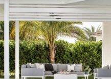 A white and gray outdoor sectional with white fire pit sits under a white pergola next to an in ground pool.