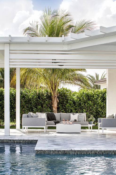 A white and gray outdoor sectional with white fire pit sits under a white pergola next to an in ground pool.