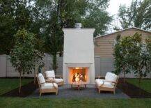 Teak chairs and a teak sofa with white cushions sits in front of a large outdoor fireplace.