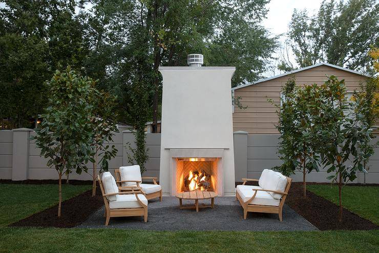 Teak chairs and a teak sofa with white cushions sits in front of a large outdoor fireplace.