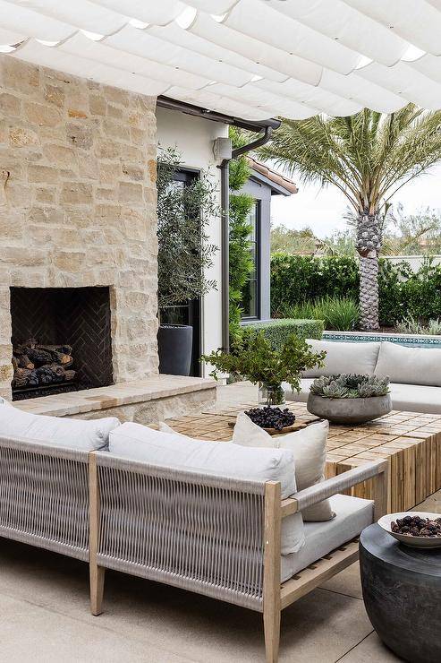 A white fabric pergola sits over an outdoor living space that features wood and gray rope outdoor sofas with a reclaimed wood coffee table, and a stone fireplace and hearth.