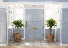 Gorgeous entryway features a blue front door matched with leaded glass sidelights. The door opens to a taupe and blue mosaic floor tiles complementing walls clad in blue millwork. The door is flanked by two potted plants in woven baskets.