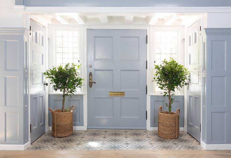 Gorgeous entryway features a blue front door matched with leaded glass sidelights. The door opens to a taupe and blue mosaic floor tiles complementing walls clad in blue millwork. The door is flanked by two potted plants in woven baskets.