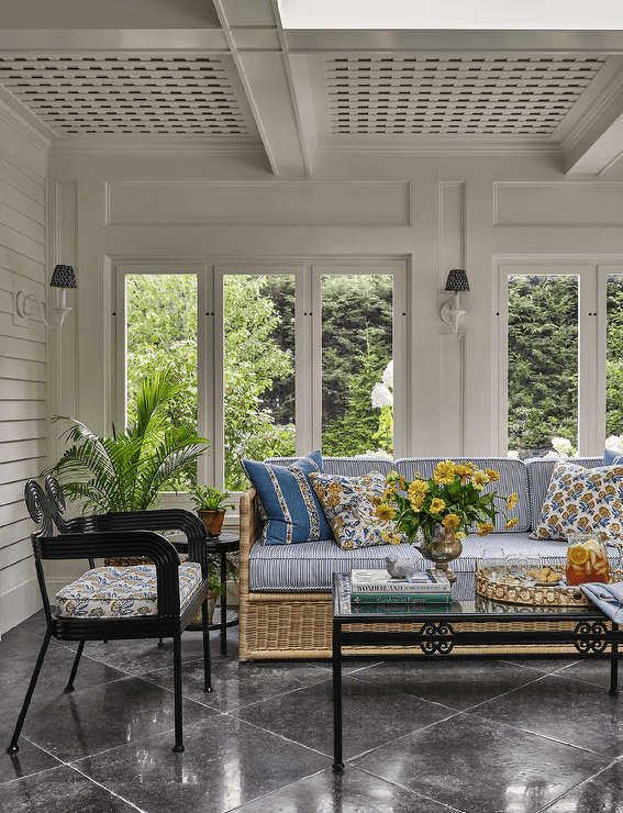 Sunroom features a rattan sofa with blue striped cushions accented with yellow floral pillows, a wrought iron coffee table and a glossy black accent chair atop black diamond pattern floor tiles under a lattice ceiling.