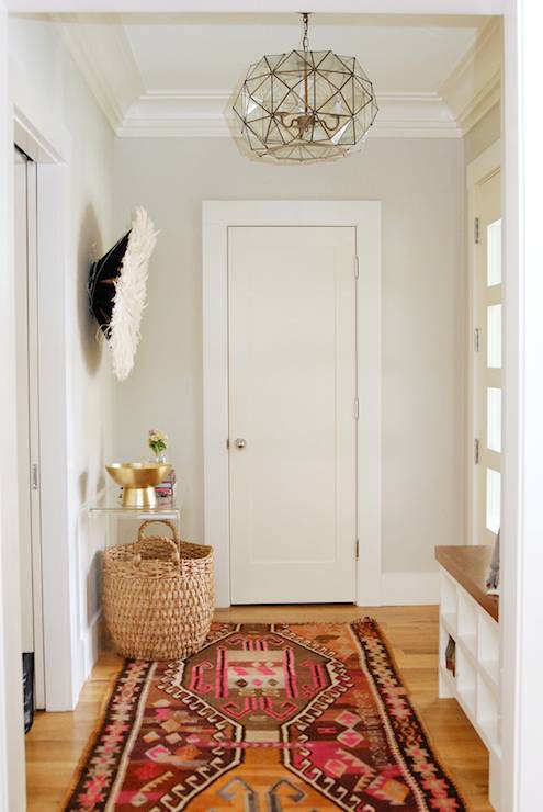 Boho foyer features greige walls over honey oak hardwood floors layered with a colorful Kilim rug illuminated by a Worlds Away Rozz Clear Pendant over a Juju Headdress and CB2 Peekaboo Clear Console Table topped with an Arteriors Acton Large Centerpiece.