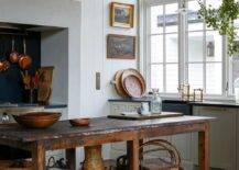 A rustic freestanding kitchen island sits in a farmhouse kitchen to the side of light gray shaker cabinets adorned with antique brass hardware topped with a black marble countertop. The countertop is fitted with a black marble apron sink with a polished brass hook and spout faucet fitted in front of a window framed by a light gray vertical shiplap trim.