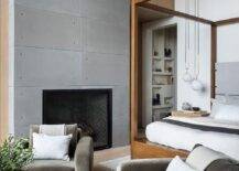 Bedroom features a sitting area with dark gray velvet accent chairs at at round black and white coffee table atop a gray rug, a modern fireplace and a wooden canopy bed under vaulted ceiling.