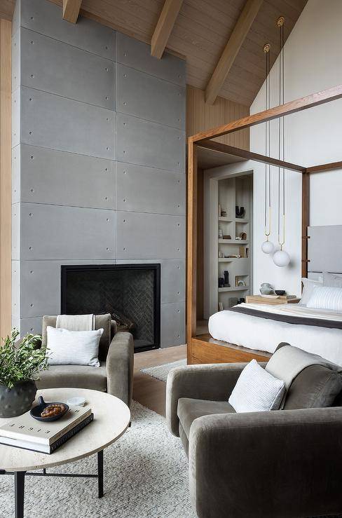 Bedroom features a sitting area with dark gray velvet accent chairs at at round black and white coffee table atop a gray rug, a modern fireplace and a wooden canopy bed under vaulted ceiling.