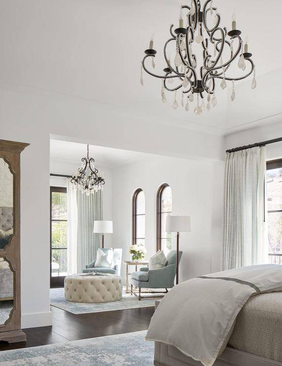 Master bedroom features a master sitting area with French crystal chandelier over gray accent chairs with a round cream tufted ottoman as coffee table atop a vintage rug.