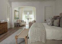 Bedroom features a gray linen bed accented with gray linen pillows, a tan bench at the foot of the bed atop a tan rug, a wooden dresser and a sitting area with cream chairs.