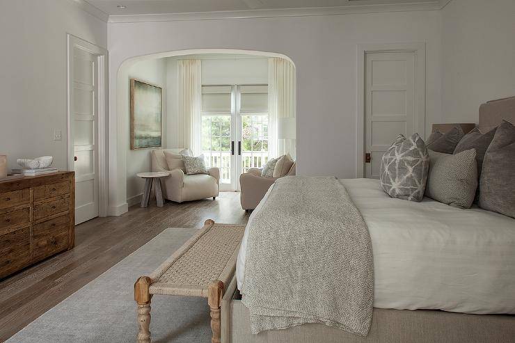 Bedroom features a gray linen bed accented with gray linen pillows, a tan bench at the foot of the bed atop a tan rug, a wooden dresser and a sitting area with cream chairs.