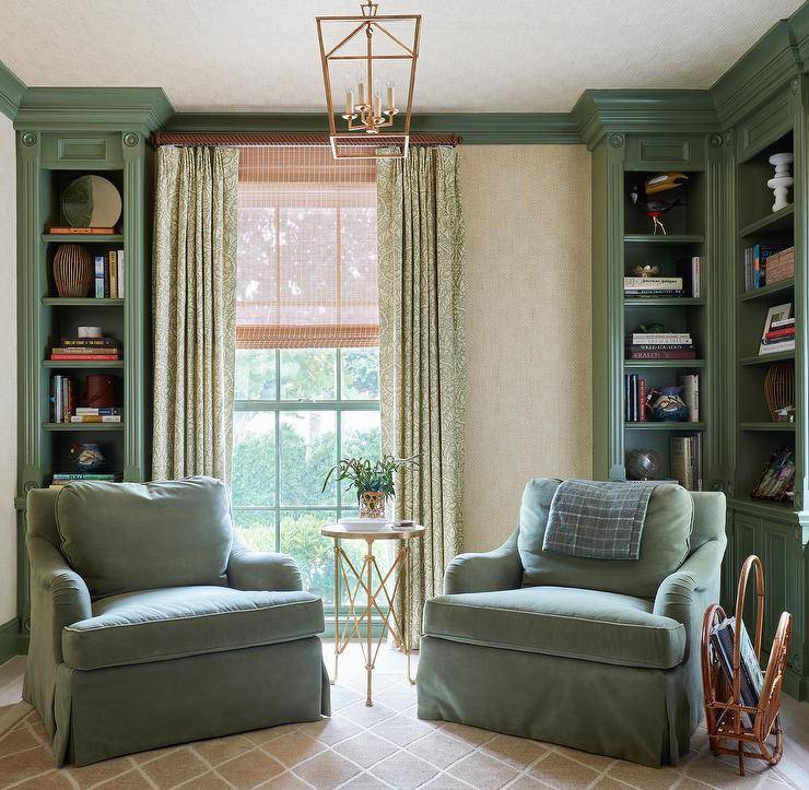 Green velvet roll arm chairs with a brass accordion table and wicker magazine holder sit on a beige lattice rug near green built in bookcases and green curtains lit by a brass lantern.