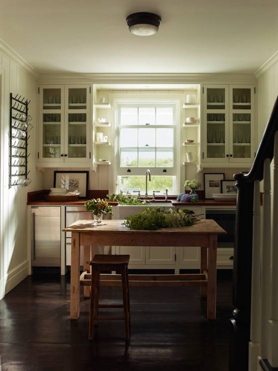 Lovely cottage kitchen with glass-front upper cabinets and white shaker lower cabinets paired with wood countertops and paneled backsplash. Oil-rubbed bronze flushmount over farmhouse kitchen island lined with rustic bar stool over wood floors. Kitchen features farmhouse sink under double hung window next to stainless steel stacked stacked dishwashers and mini-fridge.