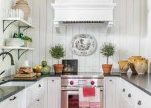 Small, charming u-shaped kitchen is equipped with a mini Wolf range placed beneath a white wooden range hood mounted to a vertical shiplap trim and behind a bright red and black rug. The range is flanked by white cabinets contrasted with oil rubbed bronze cup pulls and a black marble countertop. A farmhouse sink with an oil rubbed bronze faucet is positioned beneath a window located beside stacked white shelves.