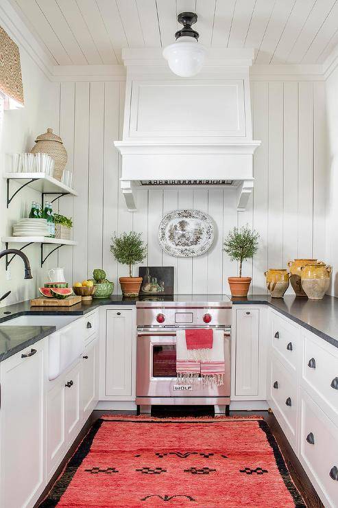 Small, charming u-shaped kitchen is equipped with a mini Wolf range placed beneath a white wooden range hood mounted to a vertical shiplap trim and behind a bright red and black rug. The range is flanked by white cabinets contrasted with oil rubbed bronze cup pulls and a black marble countertop. A farmhouse sink with an oil rubbed bronze faucet is positioned beneath a window located beside stacked white shelves.