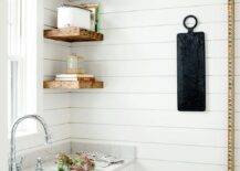 Stacked rustic wooden floating shelves are mounted against a shiplap backsplash over white kitchen drawers contrasted with oil rubbed bronze pulls. A farmhouse sink with a chrome gooseneck faucet is mounted beneath a window in a cottage-style pantry.