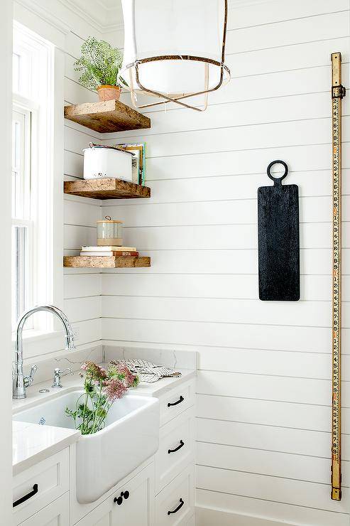 Stacked rustic wooden floating shelves are mounted against a shiplap backsplash over white kitchen drawers contrasted with oil rubbed bronze pulls. A farmhouse sink with a chrome gooseneck faucet is mounted beneath a window in a cottage-style pantry.