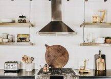 A rustic plank ceiling with rustic wood beams is mounted over a black and white kitchen featuring a stainless steel hood fixed to shiplap trim between wood and wire shelves. The shelves hang over black cabinets donning brass pulls and a black marble countertop flanking a stainless steel oven range.