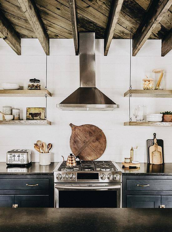 A rustic plank ceiling with rustic wood beams is mounted over a black and white kitchen featuring a stainless steel hood fixed to shiplap trim between wood and wire shelves. The shelves hang over black cabinets donning brass pulls and a black marble countertop flanking a stainless steel oven range.