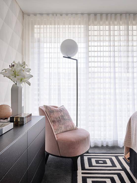 Modern bedroom features a pink velvet chair on a black and white geometric rug, a black dresser and white curtains, lit by a black and white floor lamp.