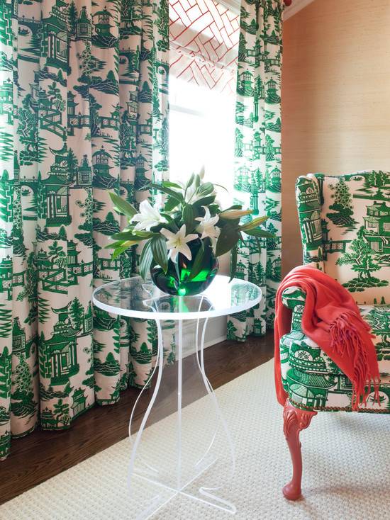 Delightful bedroom seating nook with soft coral grasscloth wallpaper as the perfect backdrop to incredible emerald green and white Chinoiserie fabric in floor-length drapes and also upholstered upon an updated wingchair. Lovely partnering with Simone Mirrored Table and emerald green vase as contrast to additional color in a lovely coral throw. Balanced flooring in a beautiful cream rug.